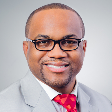 A smiling man wearing black glasses, a light gray suit, a white shirt, and a red patterned tie, posing against a plain light gray background.