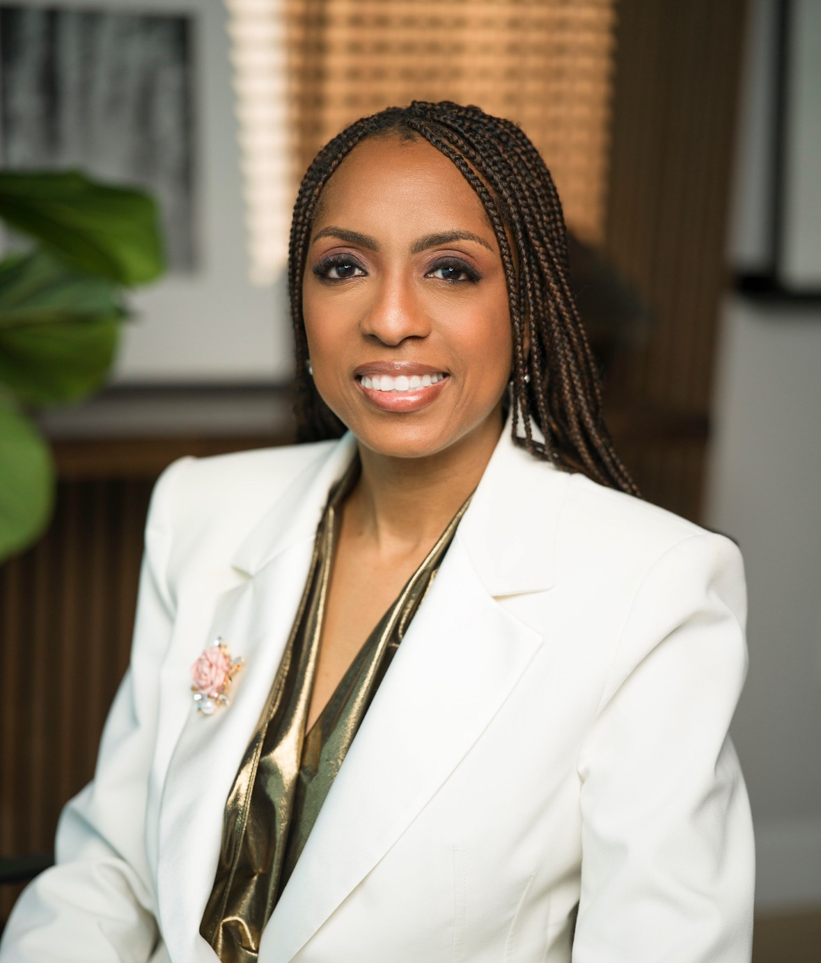 A woman with braided hair, wearing a white blazer with a brooch and a shiny gold top, sits smiling in an indoor setting with blurred background decor.